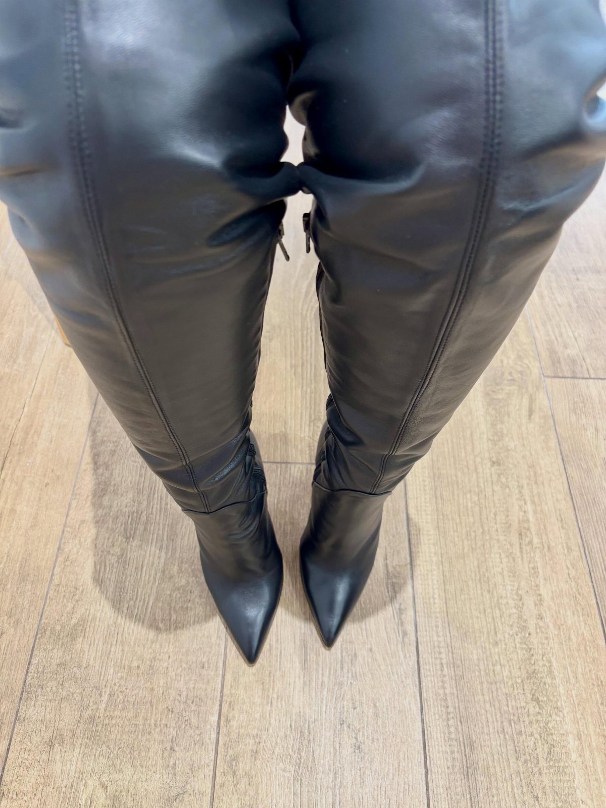 Black over-the-knee pointed-toe boots worn on a light wooden floor.