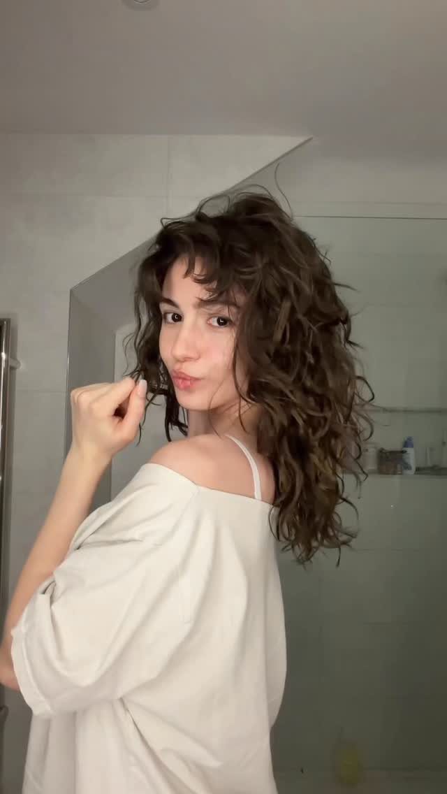 Woman with long brown wavy hair posing in a white tiled bathroom