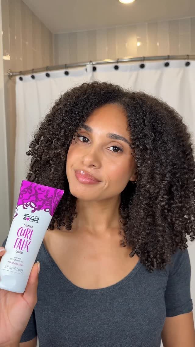 Woman with curly hair holding a tube of curl defining cream in a bathroom
