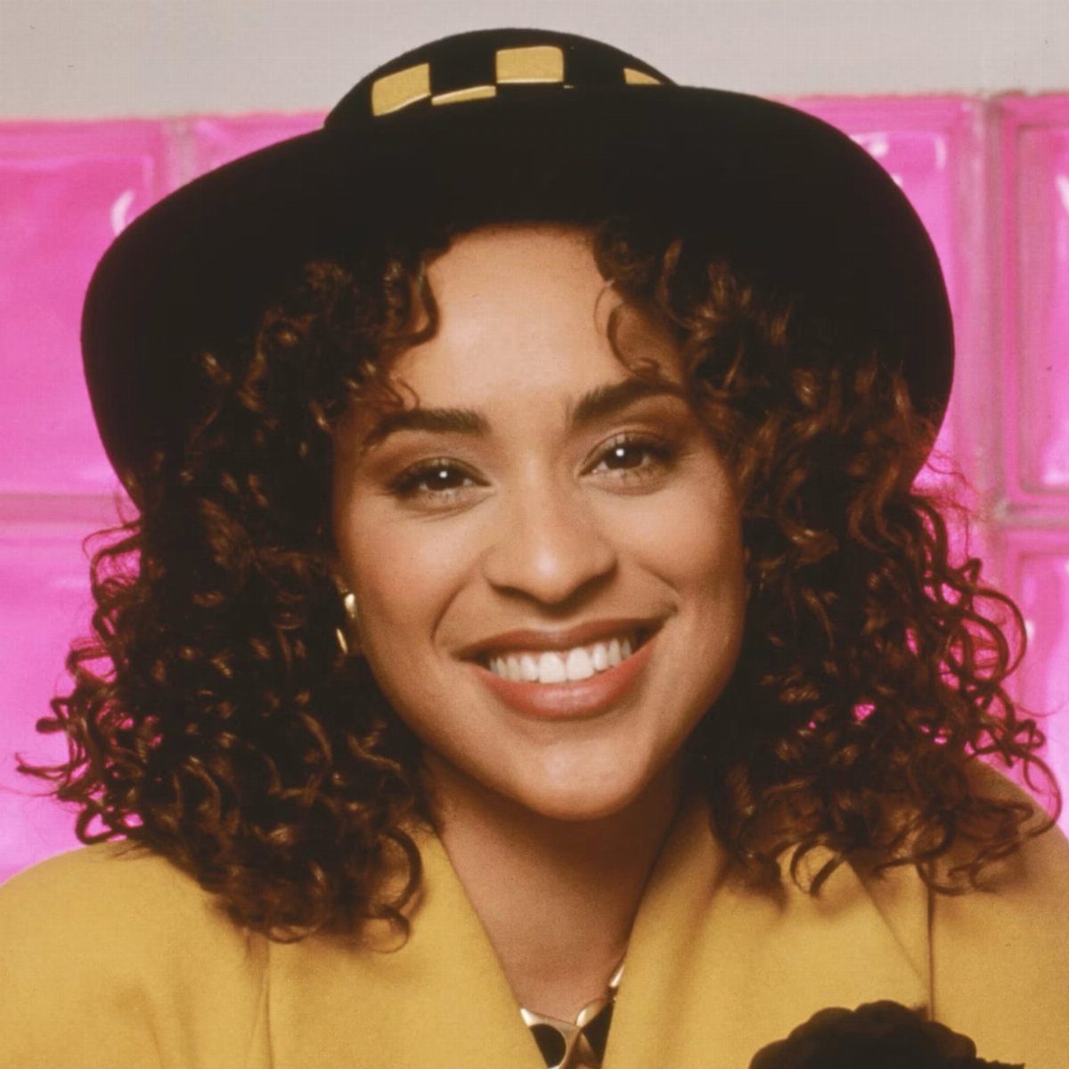 Woman with voluminous curly brown hair wearing a black hat and yellow blazer.