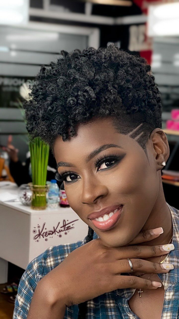 Woman with a short curly black pixie cut and shaved side designs smiling.