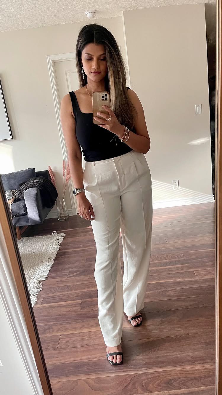 Woman in a black top and wide-leg white trousers taking a mirror selfie.