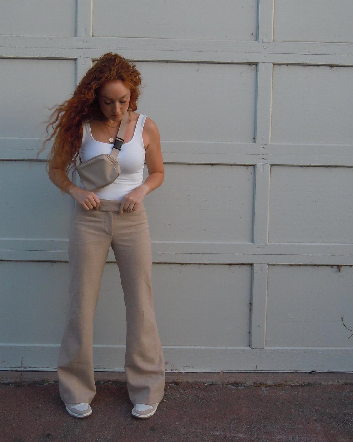 Woman wearing beige wide-leg trousers and a white tank top against a white wall