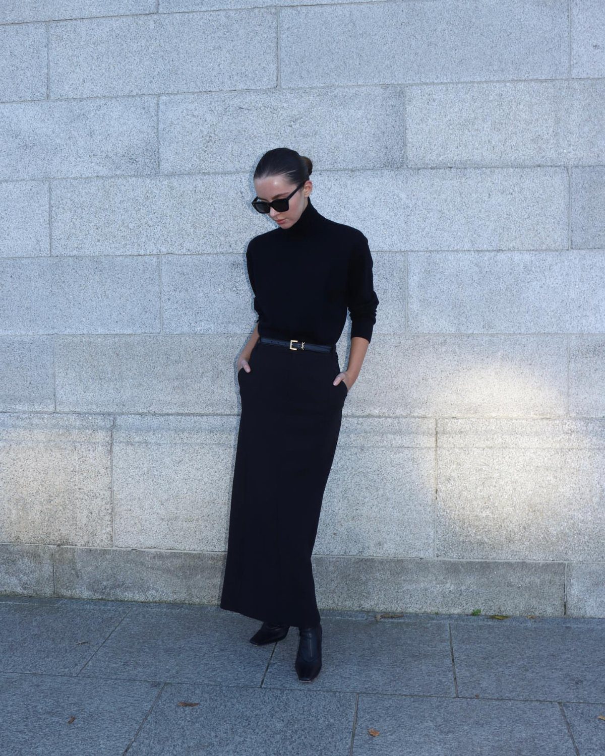 Woman in black turtleneck and black maxi skirt posing against a stone wall.