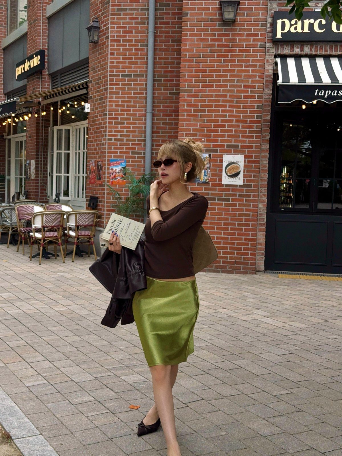 Woman wearing a brown long-sleeved top and olive green satin midi skirt outdoors.