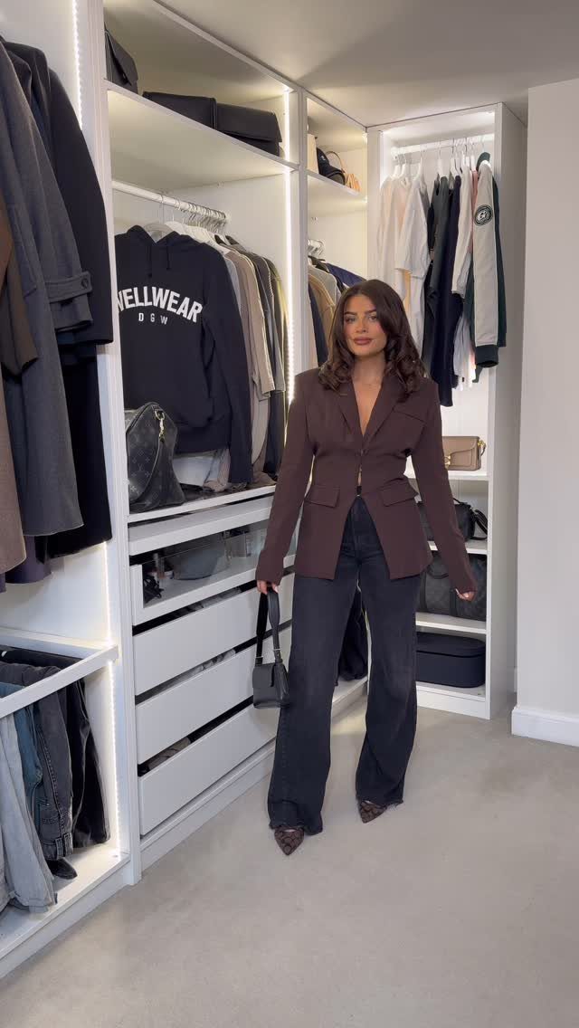 Woman in a brown blazer and grey trousers posing in a white closet.