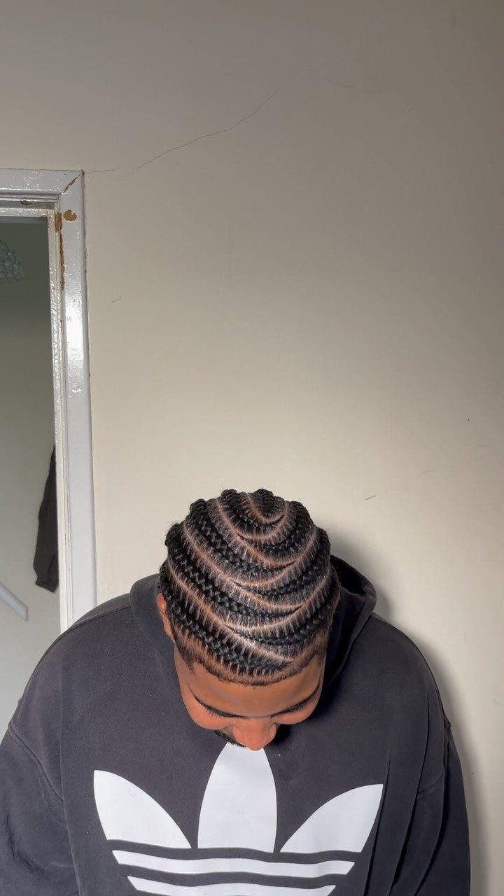 Man with curved cornrow braids wearing a black Adidas hoodie against a wall