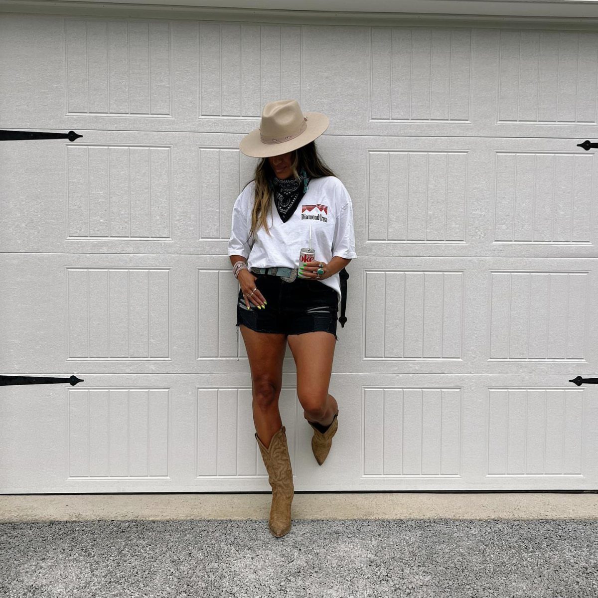 Woman in white graphic tee, black shorts, cowboy hat, and tan Western boots.