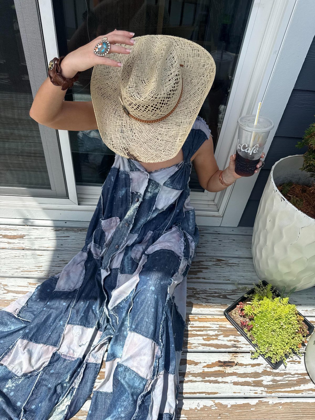 Person in a patchwork denim dress and straw hat sitting on a deck.