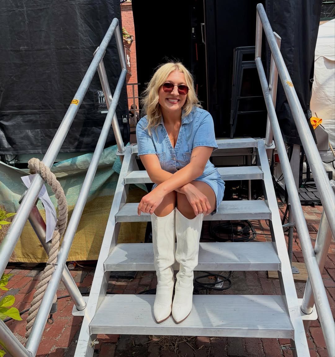 Woman in a blue denim romper and white cowboy boots sitting on stairs.