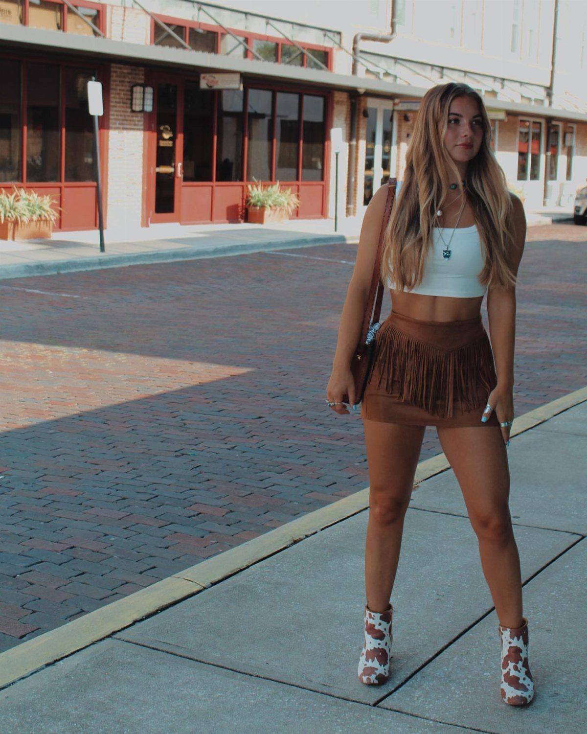 Woman wearing white crop top, brown fringed mini skirt, and patterned ankle boots.