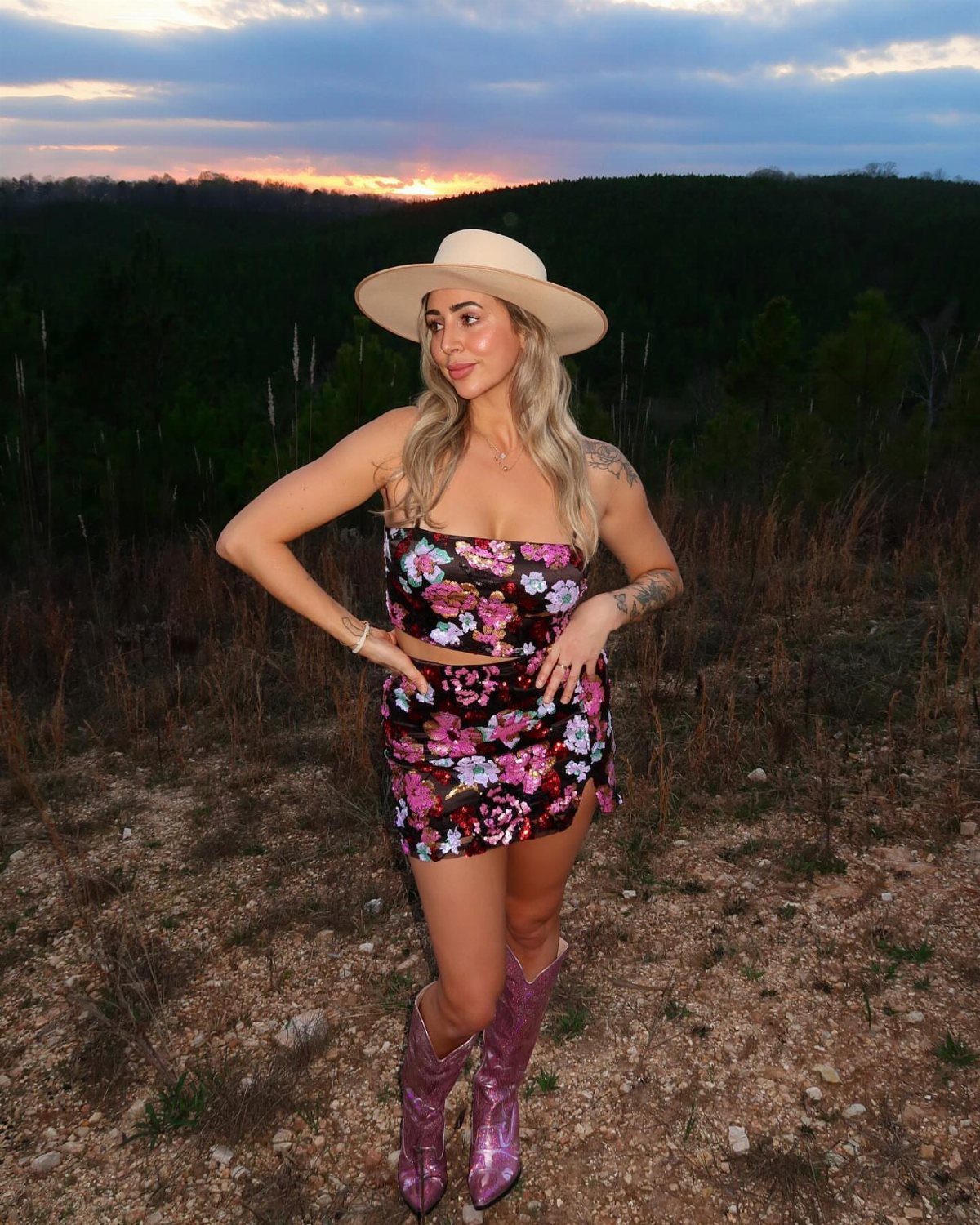Woman in floral two-piece set, purple cowboy boots, and cream hat at sunset.