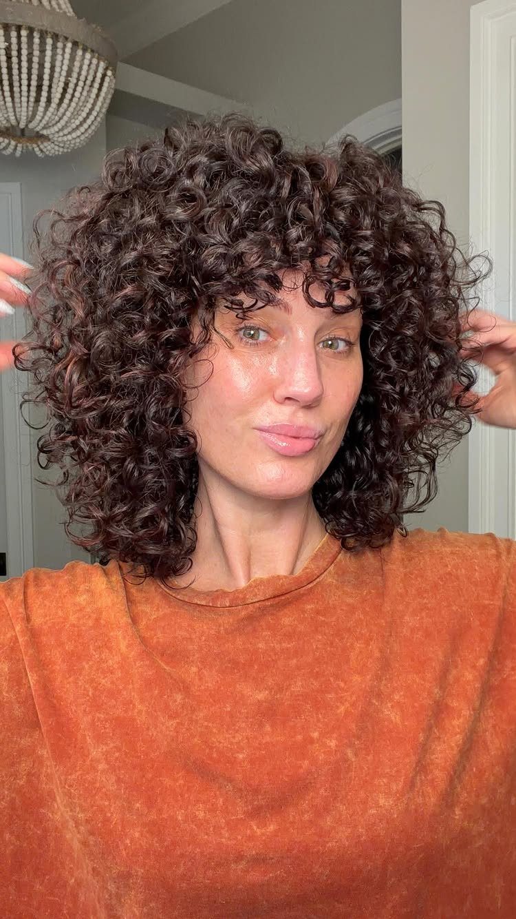Woman with voluminous dark brown curly hair and curly bangs wearing orange shirt