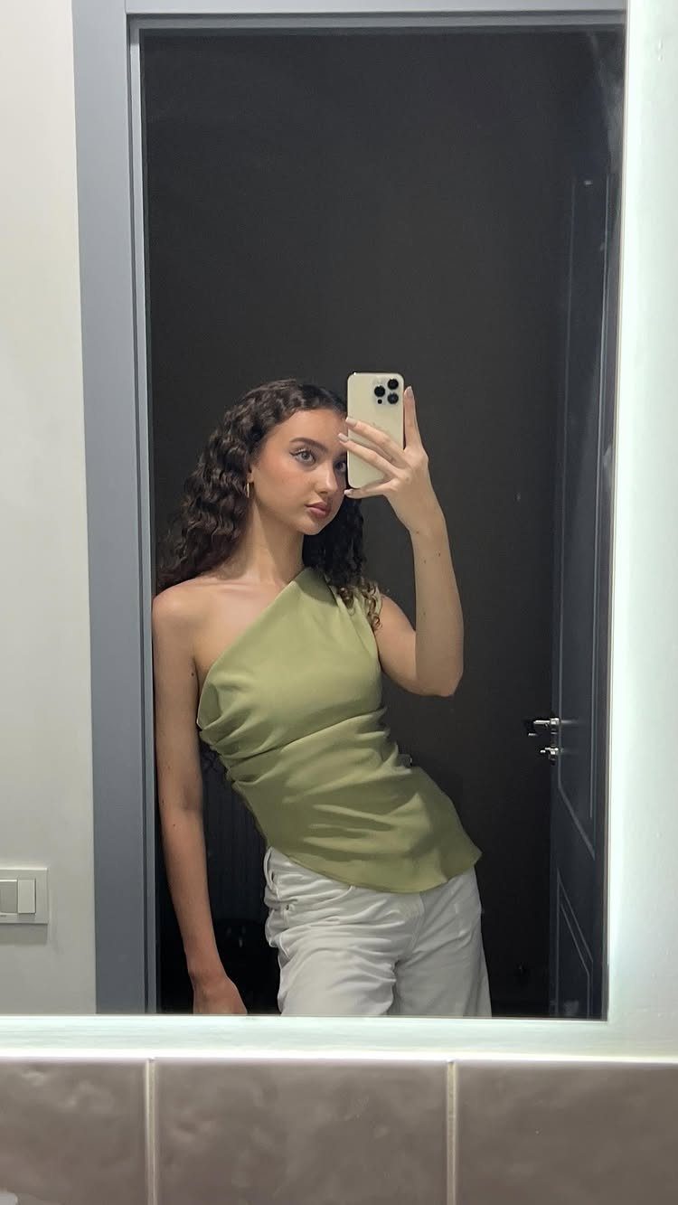 Woman with long dark curly hair wearing an olive green one-shoulder top.