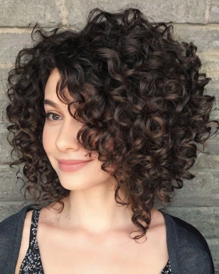 Woman with voluminous dark brown curly hair posing against a grey brick wall