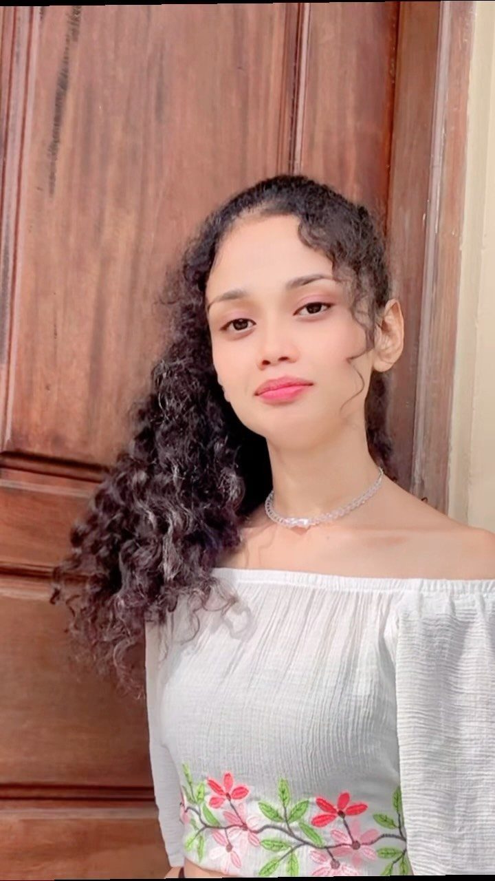 Woman with long dark curly hair wearing a white floral off-the-shoulder top.