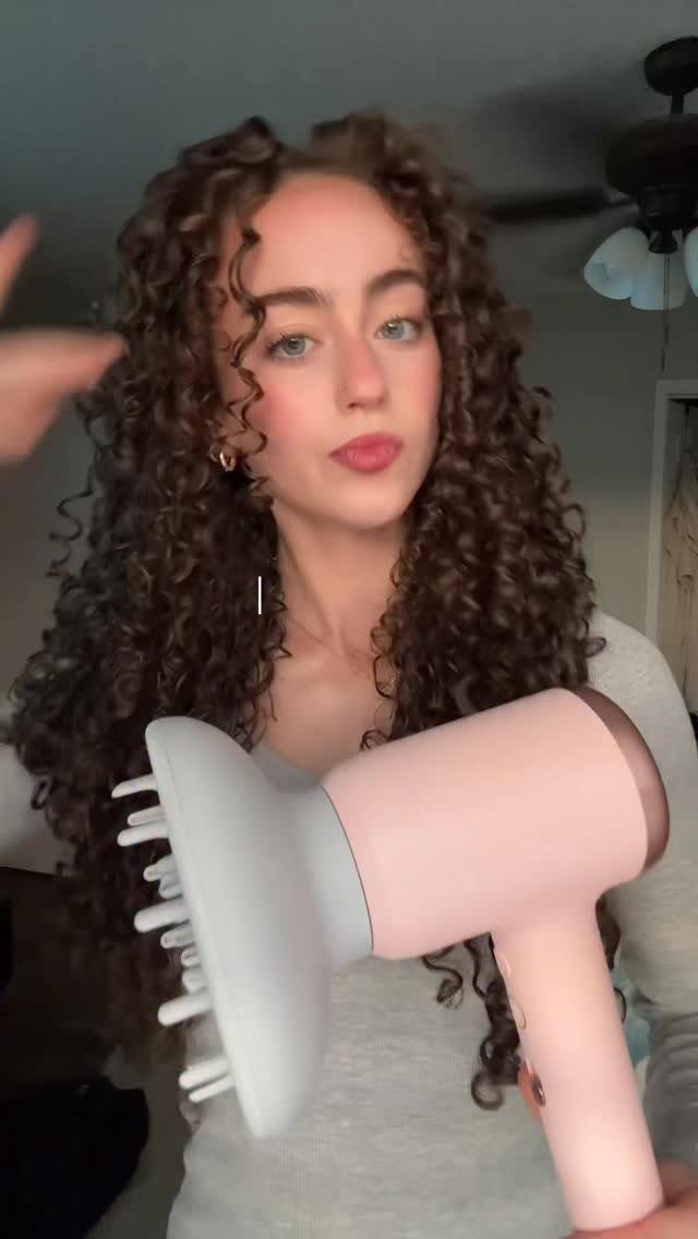 Woman with curly brown hair holding a pink hair dryer with a diffuser attachment.