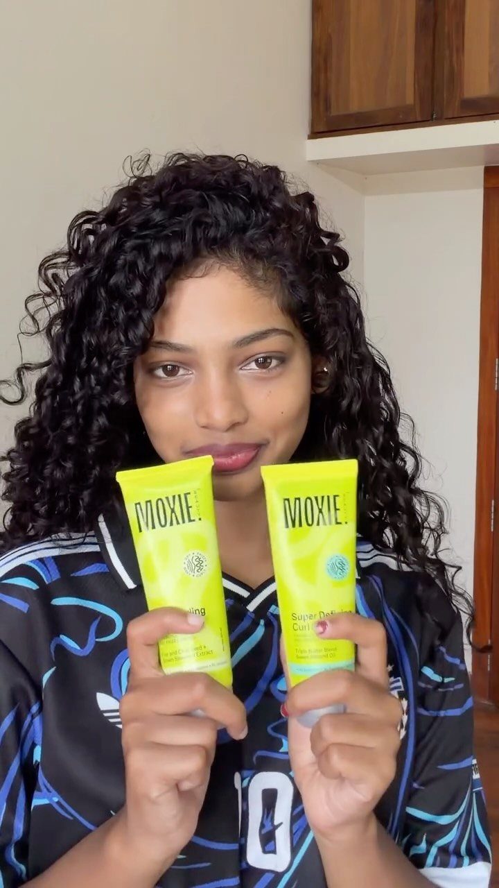 Woman with curly hair holding two yellow tubes of Moxie styling products.