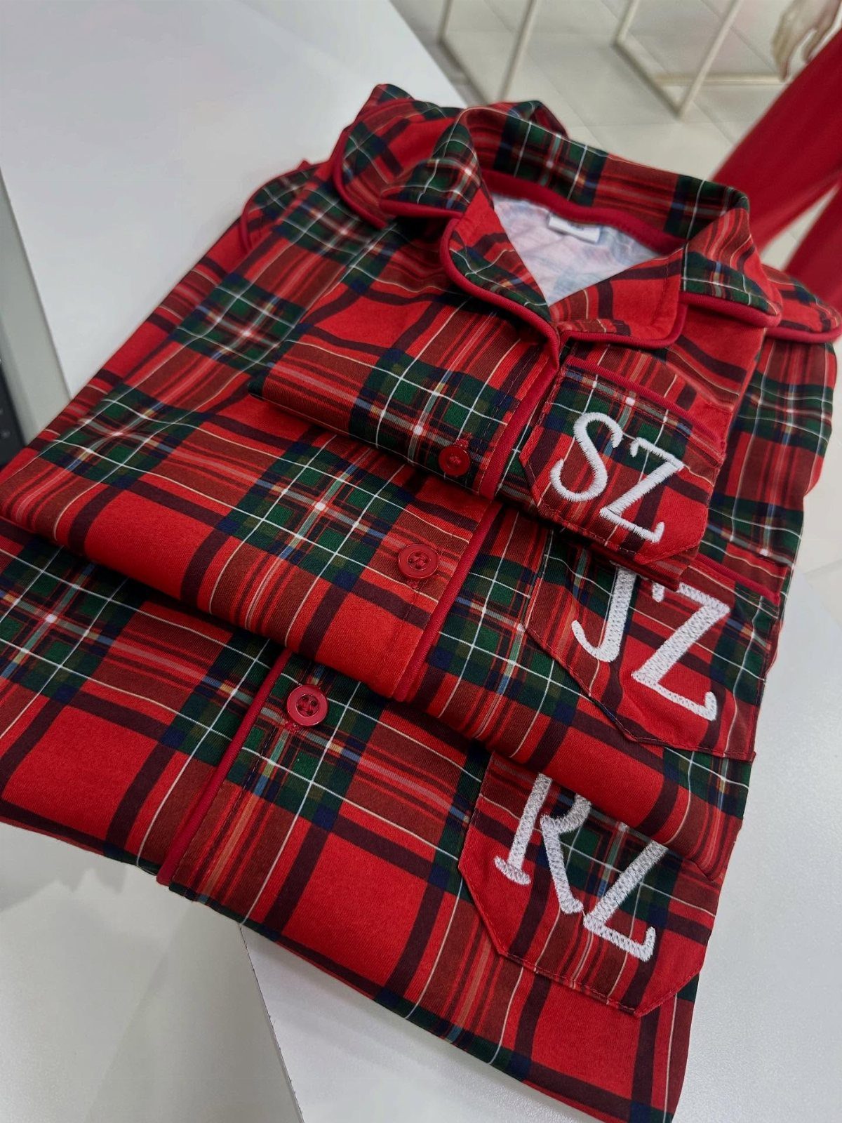 Three folded red plaid flannel pajama shirts with white embroidered initials on pockets.