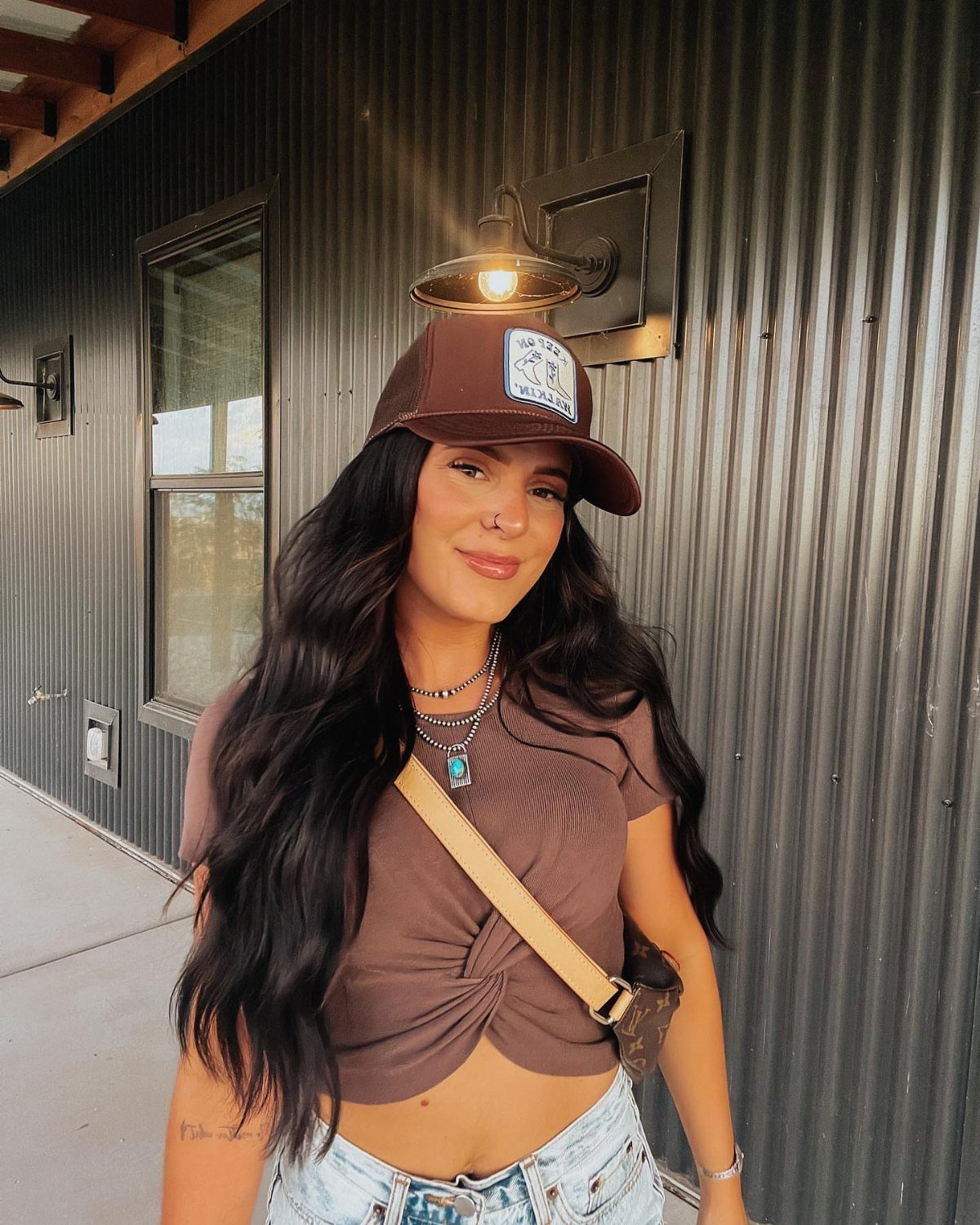 9 Cute Cowboy Outfits with Boots, Denim and Western Flair 4 Woman wearing a brown trucker hat, brown cropped shirt, and light denim jeans.