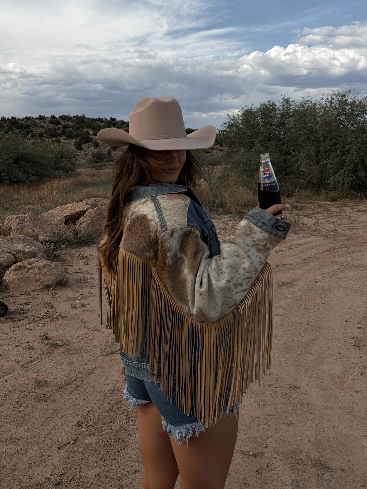 9 Cute Cowboy Outfits with Boots, Denim and Western Flair 5 Woman in a fringed western jacket and cowboy hat holding a soda bottle.