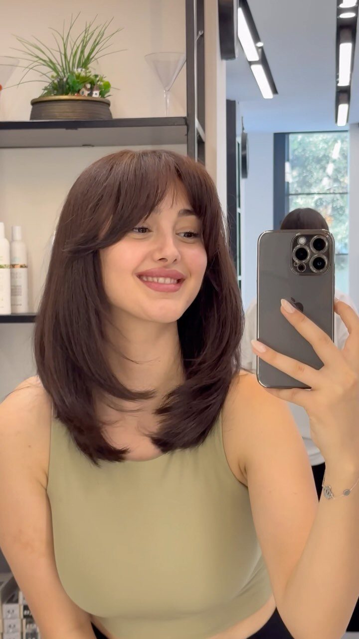 Woman with brown hair and curtain bangs taking a mirror selfie in a salon.