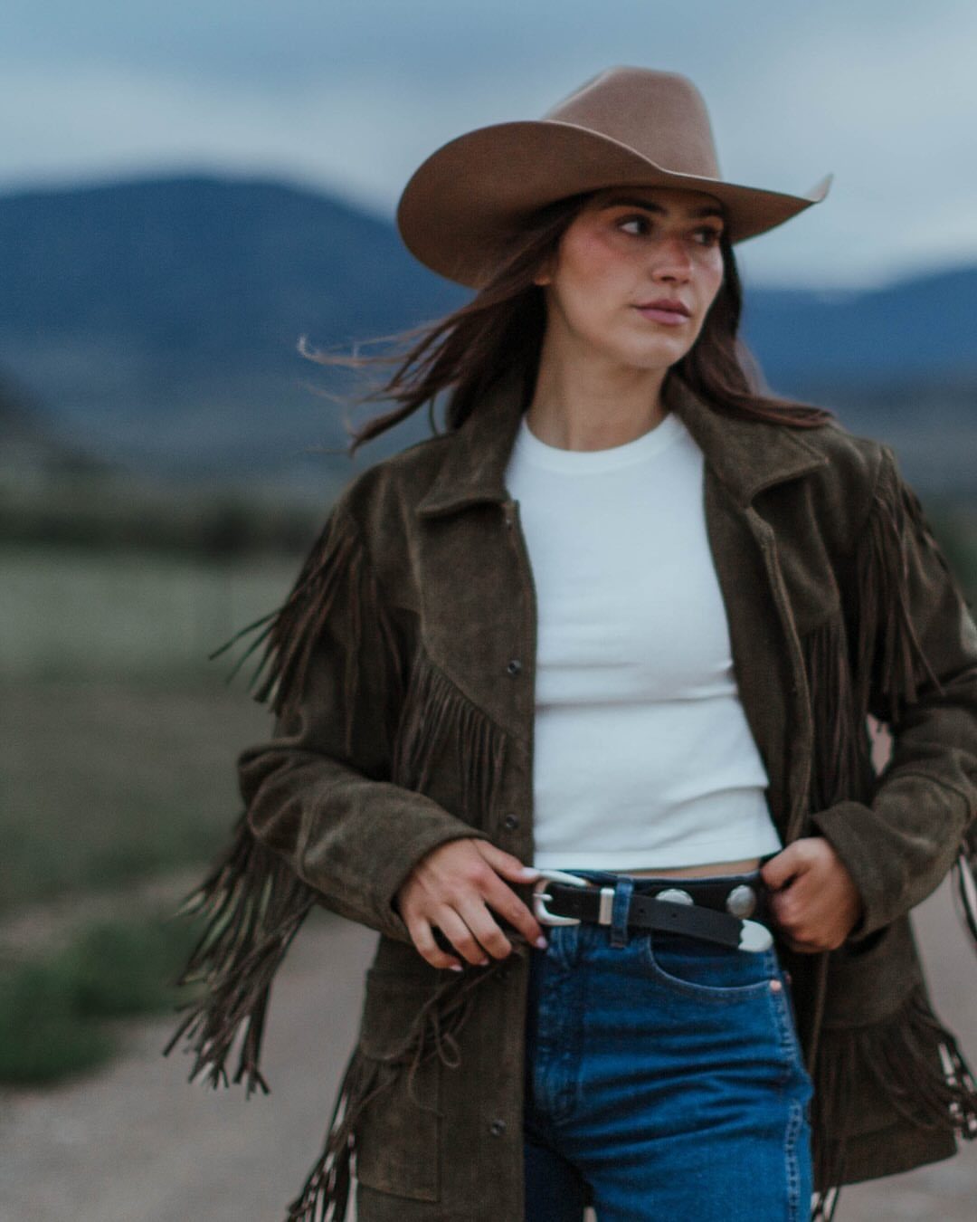 Cute Western Outfits for Women: 13 Ideas to Wear Now 7 Woman in brown fringe jacket, white shirt, blue jeans, and cowboy hat outdoors.