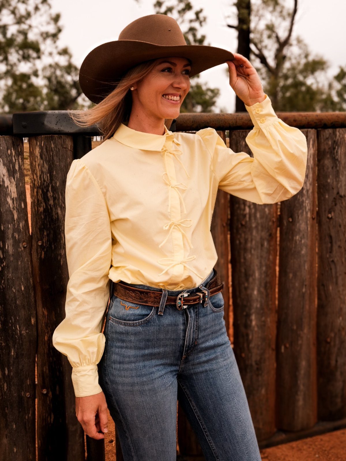 Cute Western Outfits for Women: 13 Ideas to Wear Now 2 Woman in yellow western shirt, brown cowboy hat, and blue jeans by wooden fence