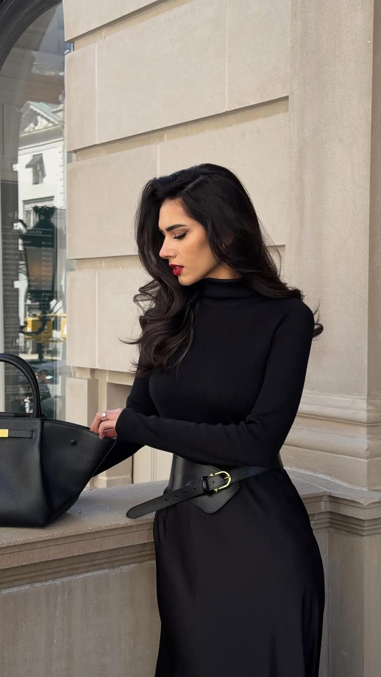 Woman in a black dress and belt posing against a stone wall.