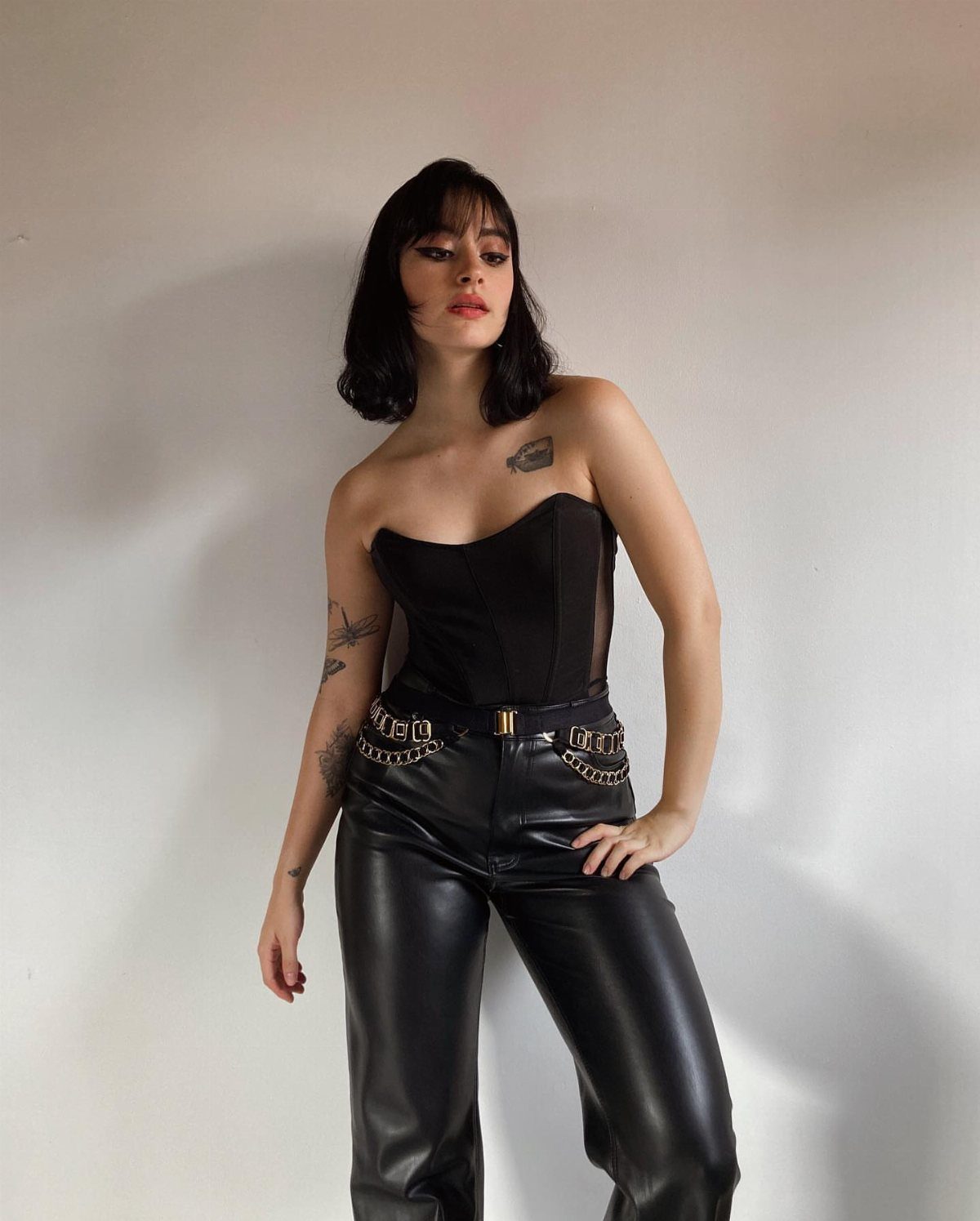 Woman in black corset and leather pants posing against a white wall.