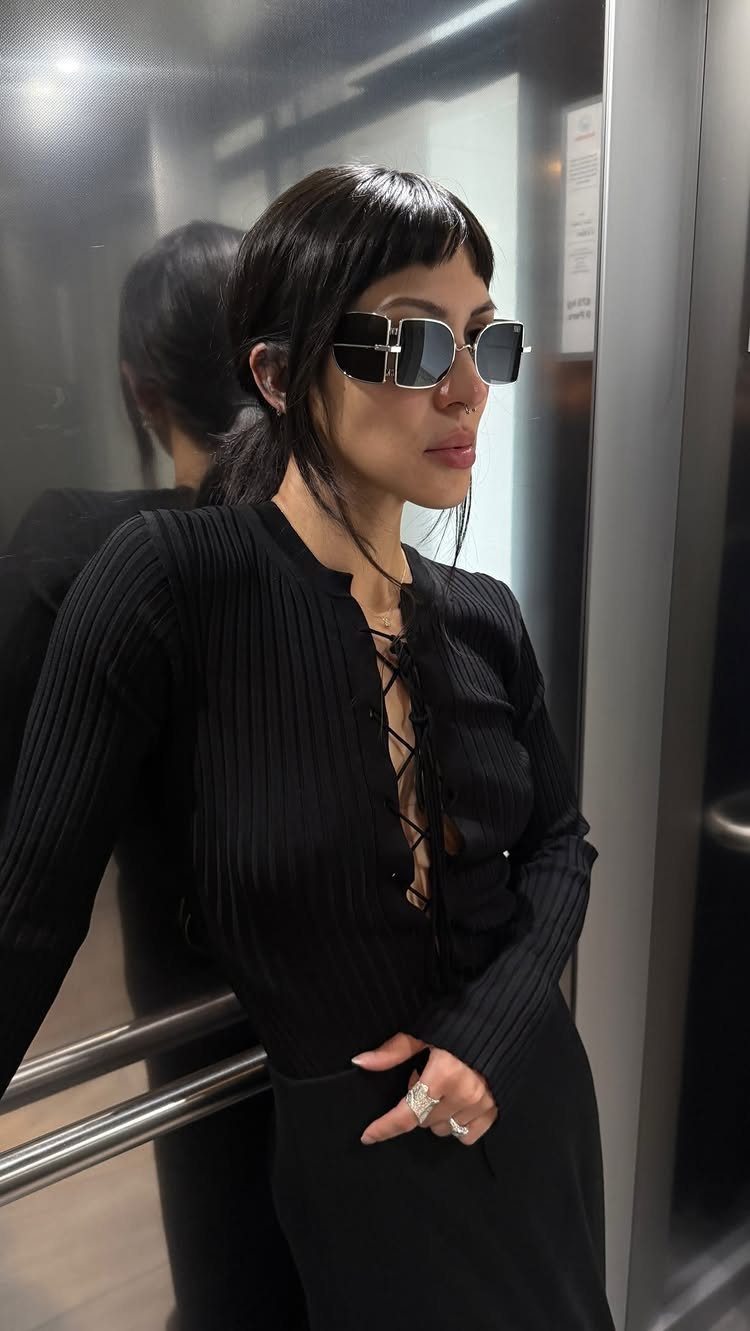 Woman in black ribbed dress and rectangular sunglasses posing in a metallic elevator