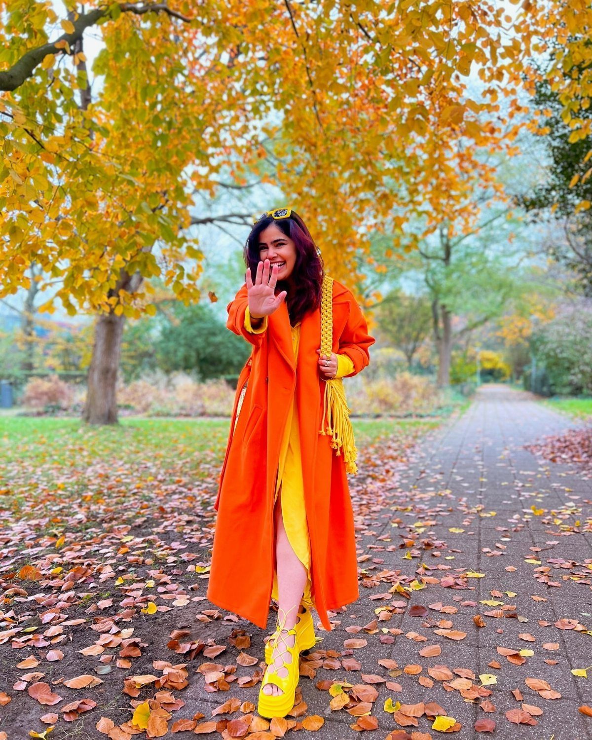 Fall Orange Outfits: 10 Ideas for Every Occasion 9 Woman in bright orange coat and yellow dress standing on autumn leaf path