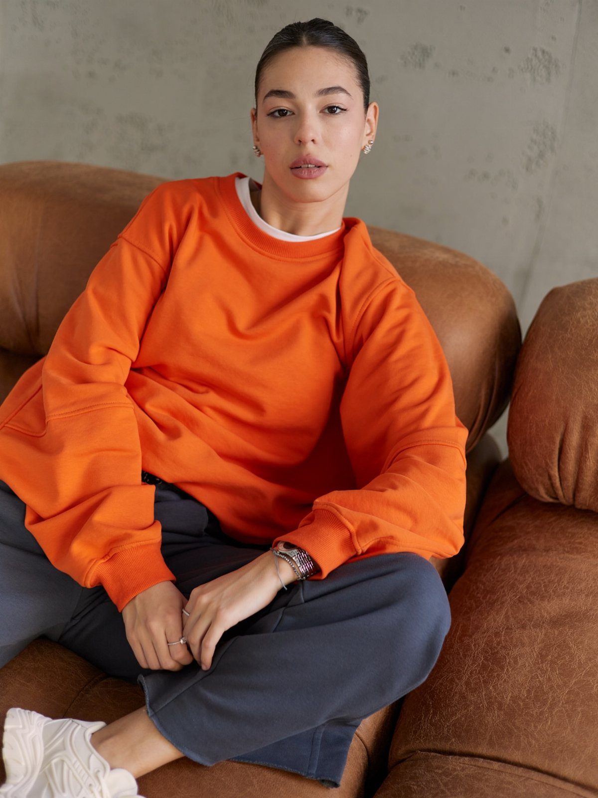 Fall Orange Outfits: 10 Ideas for Every Occasion 4 Woman wearing an oversized orange sweatshirt and grey trousers sitting on a leather sofa.