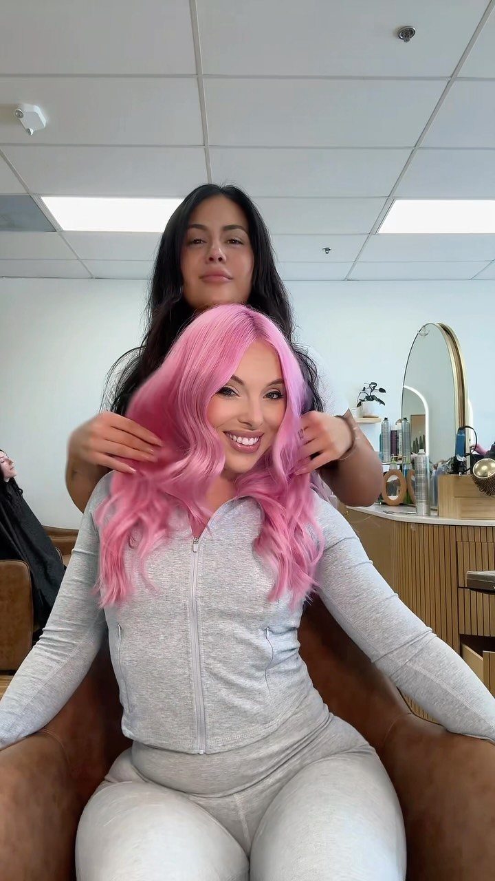 Woman with shiny, wavy pink hair sitting in a salon chair with stylist.