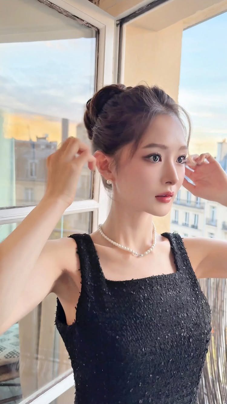 Woman with a high hair bun wearing a black dress and pearl necklace.