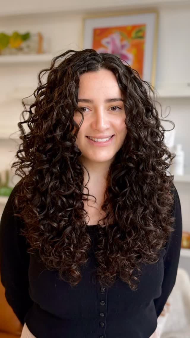 Woman with long, dark, curly hair smiling in a brightly lit room.