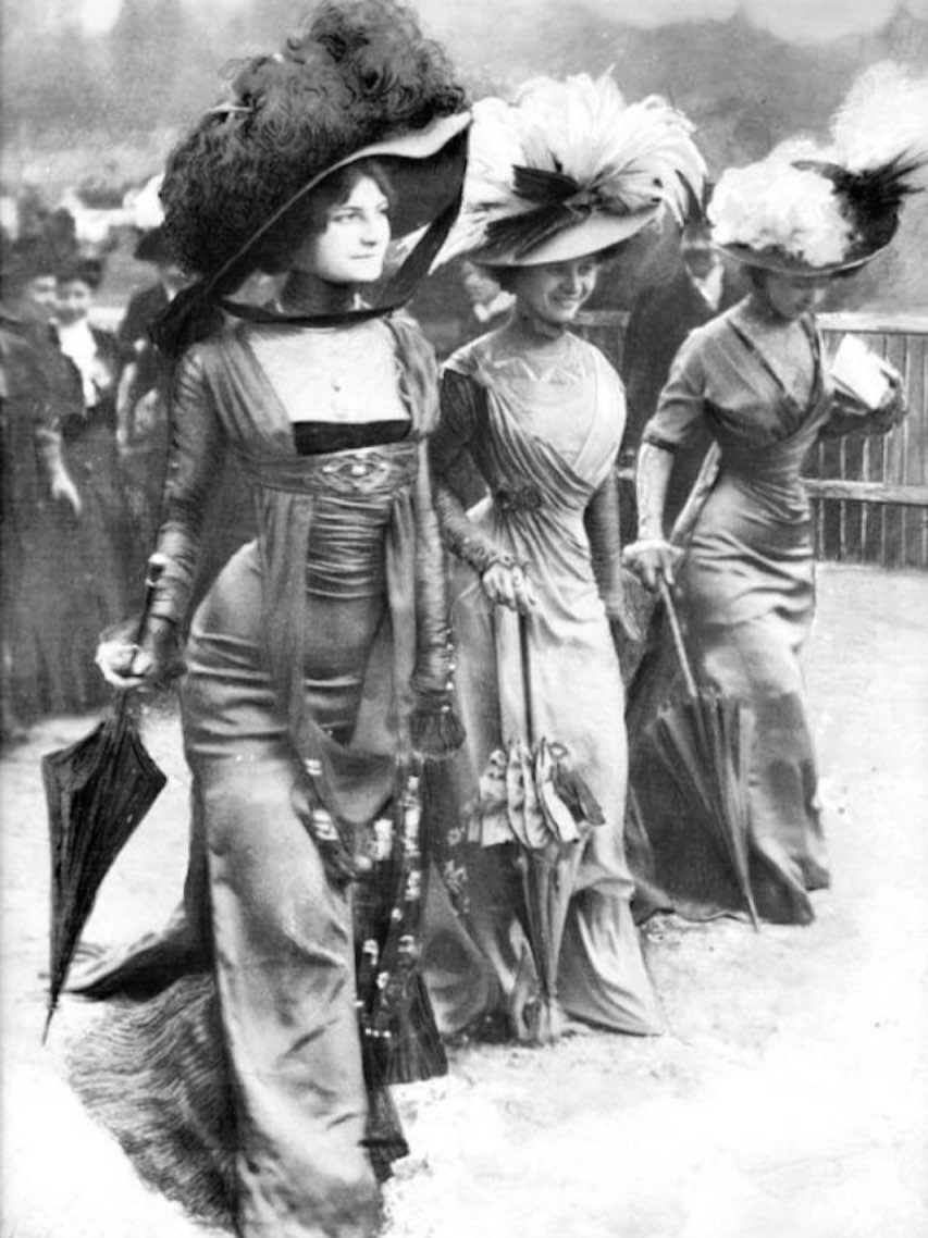 Three women in early 1900s gowns with large plumed hats and parasols.