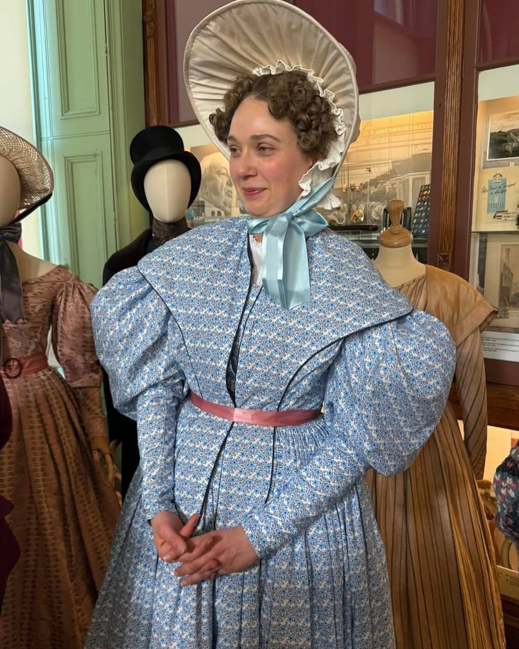Woman in a blue patterned period dress and white bonnet standing among mannequins.