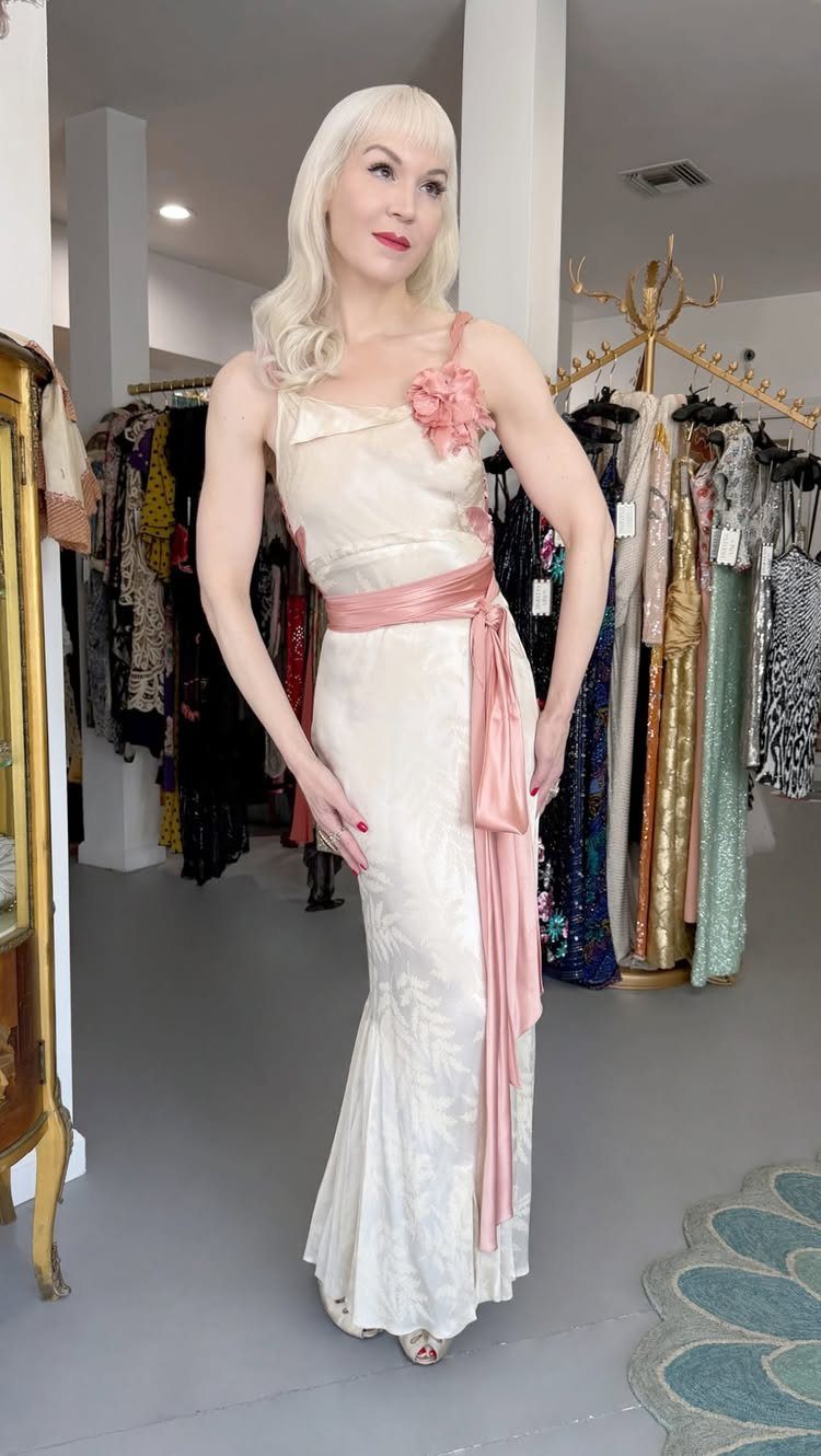 Woman in a cream satin gown with pink sash posing in a boutique.