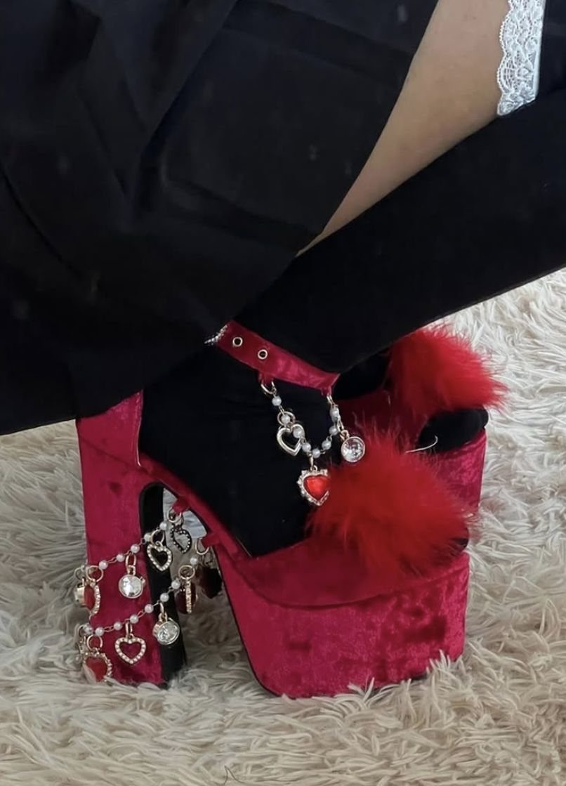 Red velvet platform heels with faux-fur pom-poms and silver heart charms on feet.