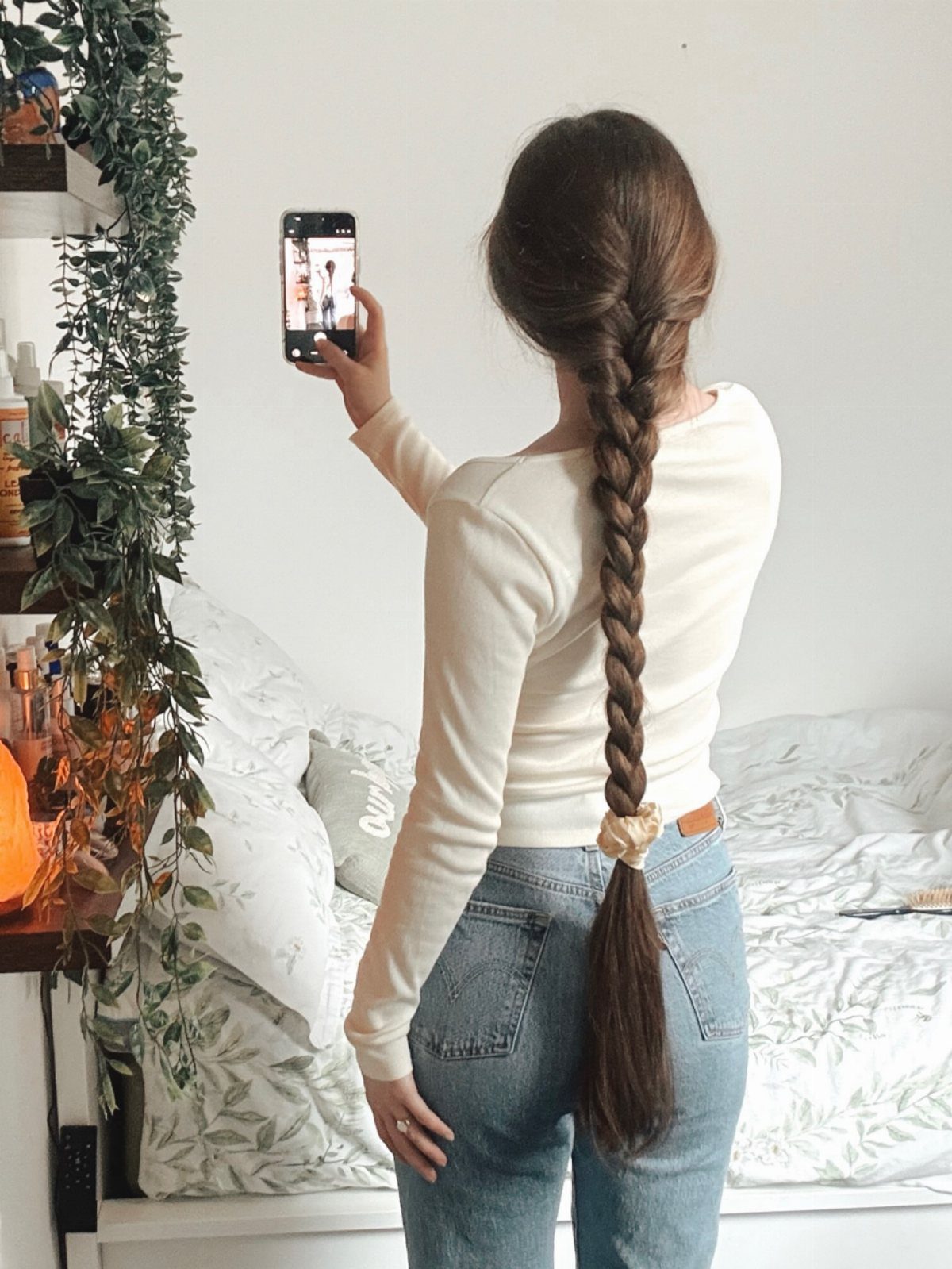 9 Long Hair Tutorial Ideas: Braids, Buns and Ponytails 4 Woman with a long braided ponytail taking a mirror selfie in her bedroom.