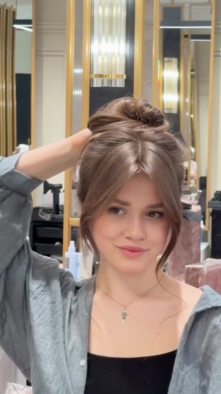 Woman with brown hair in a high messy bun and bangs in a salon.