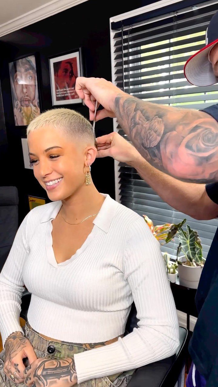 Woman with a blonde buzz cut getting a trim from a tattooed barber.