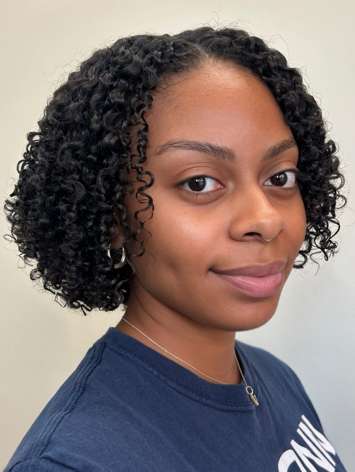 Woman with a curly bob hairstyle wearing a navy blue t-shirt and gold necklace.