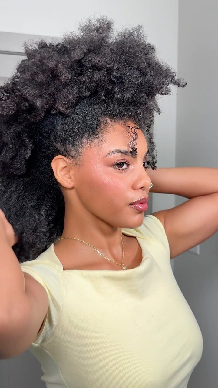 Woman with a curly frohawk hairstyle wearing a pale yellow sleeveless top.