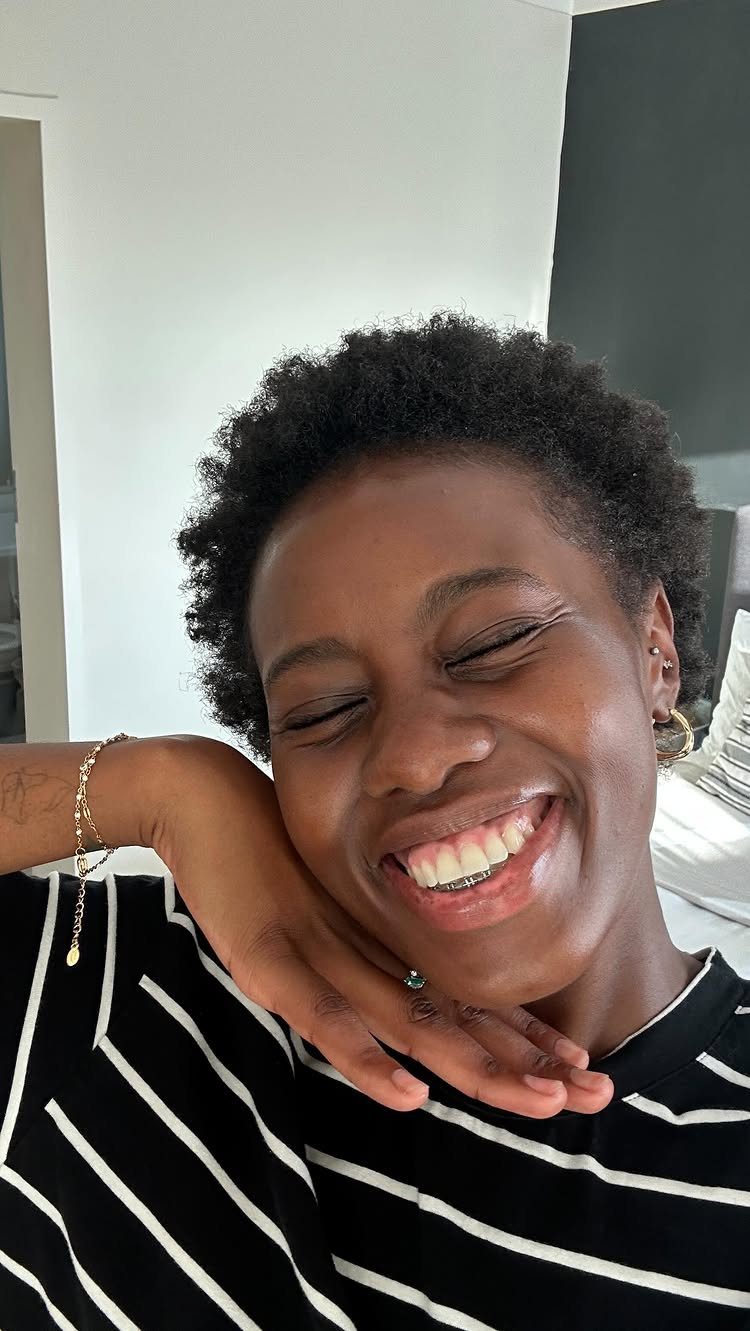 Woman with a short black afro smiling and posing against a white wall.