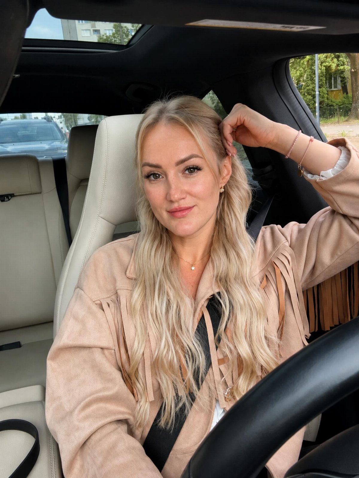 Woman with long wavy ash blonde hair wearing a tan fringed suede jacket.