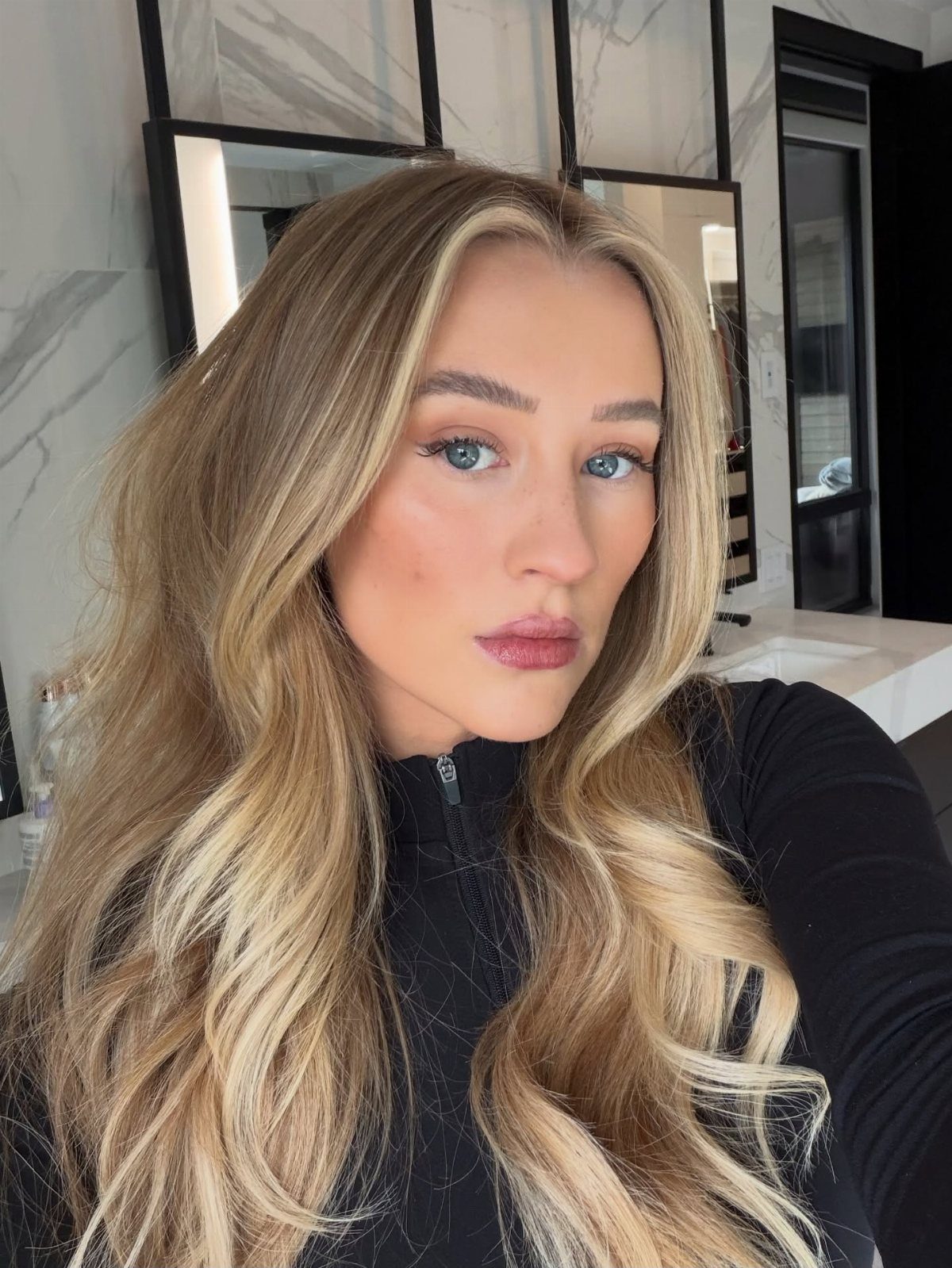 Woman with long wavy golden blonde hair posing in a marble bathroom.