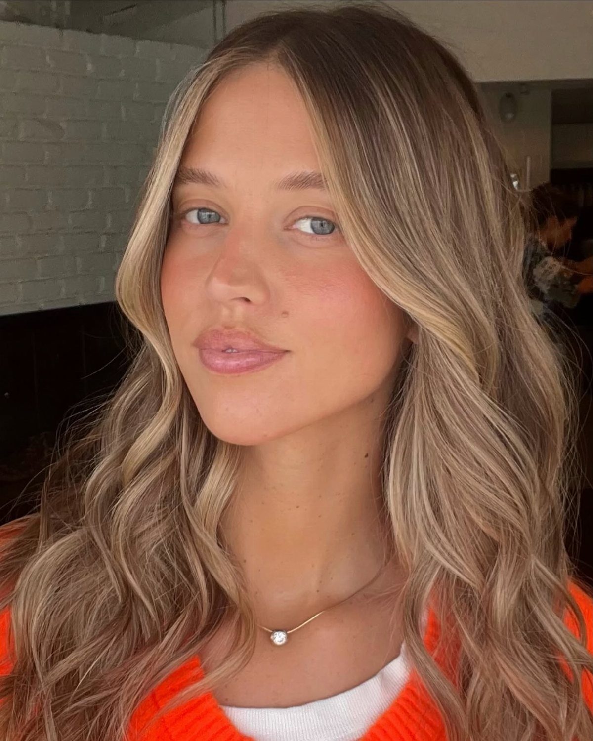 Woman with wavy blonde hair and blue eyes wearing an orange top.