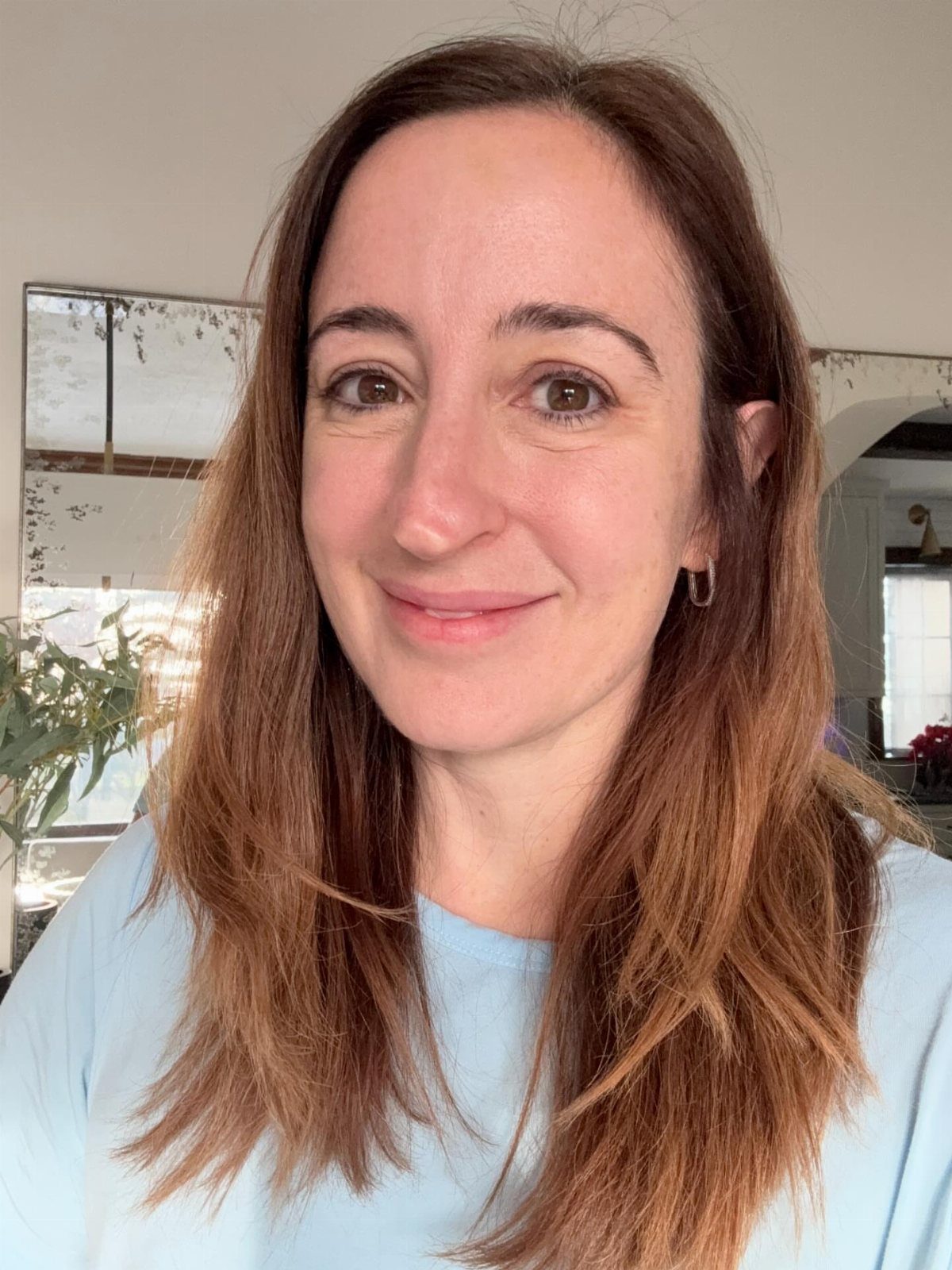 Woman with reddish-brown hair smiling, wearing a light blue shirt in a home setting.